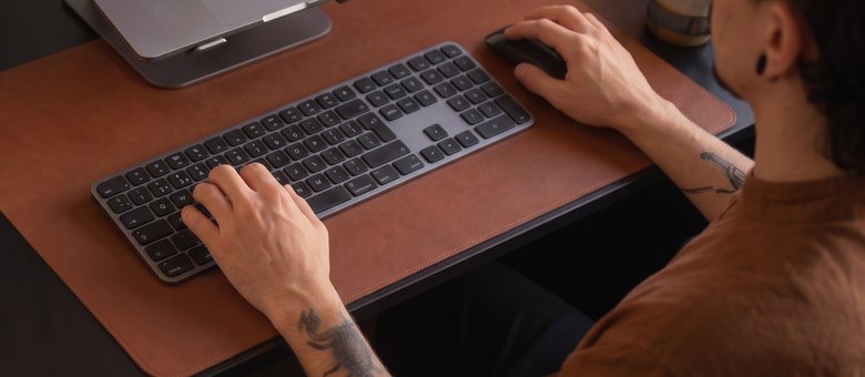 Mãos no teclado e no mouse, com o notebook apoiado atrás, na mesa de trabalho ao fim da tarde.